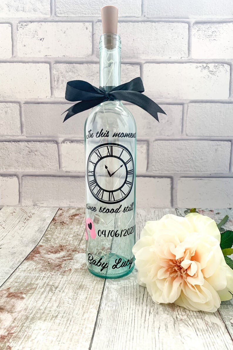 May include: A clear glass bottle with a cork top and a black ribbon tied around the neck. The bottle has a clock face with the words "In this moment time stood still" and the date "04/06/2021". The bottle also has the words "Baby Lucy" written on it.