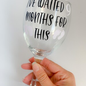 May include: A clear glass wine glass with the text "I've waited 9 months for this" and two white baby footprints on the side.