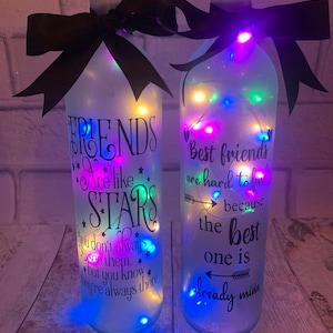 May include: Two clear glass bottles with white frosted paint and a black ribbon tied around the top. The bottles are lit with white fairy lights and have black text that reads "Friends are like stars you don't always see them, but you know they're always there" and "Best friends are hard to find because the best one is already mine".