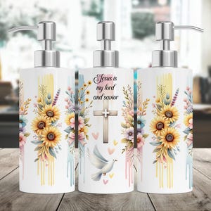 Op de afbeelding: Drie witte keramische zeepdispensers met zilveren pompen. Elke dispenser heeft een bloemmotief met zonnebloemen en de tekst "Jezus is mijn Heer en Redder" op de middelste dispenser.