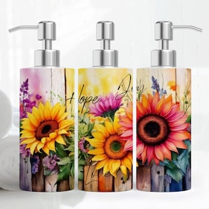 May include: Three decorative soap dispensers with floral designs. The dispensers are white with a woodgrain pattern and feature sunflowers, lavender, and other flowers. The dispensers have the words "Hope", "Joy", and "Love" written on them.