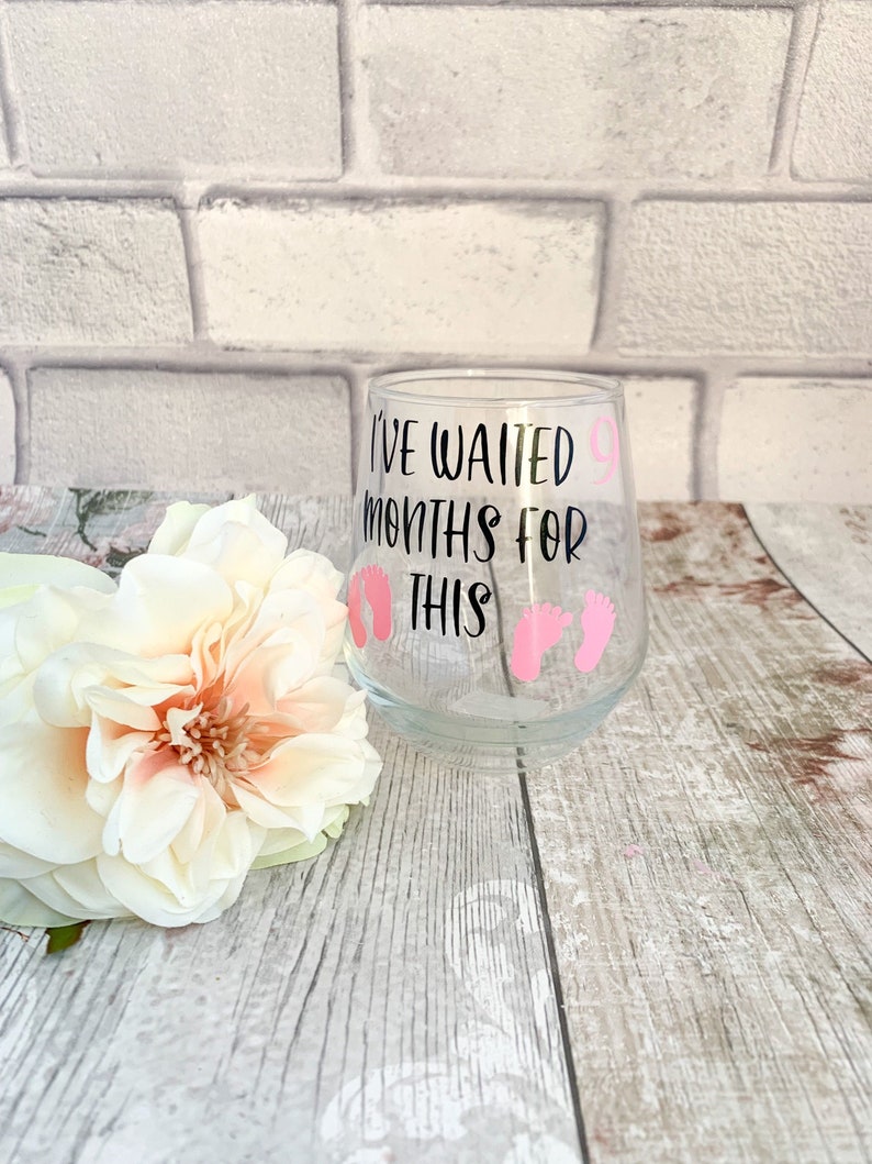 May include: A clear glass with a black and white design that says "I've waited 9 months for this" with pink baby footprints.