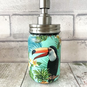 May include: A turquoise blue glass soap dispenser with a stainless steel pump and lid. The dispenser features a tropical print with a toucan, pineapple, and palm leaves.