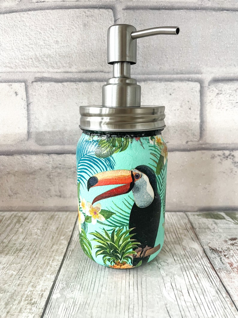 May include: A turquoise blue glass soap dispenser with a stainless steel pump and lid. The dispenser features a tropical print with a toucan, pineapple, and palm leaves.