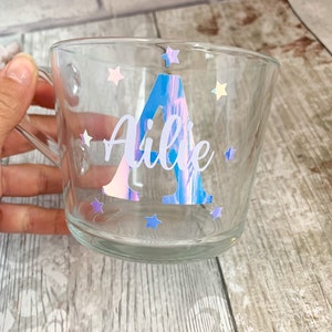 Op de afbeelding: Een heldere glazen mok met een holografische sticker met de letter "A" en de naam "Ailie" omringd door sterren.