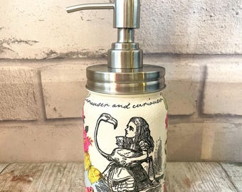 May include: A white ceramic soap dispenser with a stainless steel pump and lid. The dispenser features a black and white illustration of Alice in Wonderland with a flamingo. The text "curiouser and curiouser" is printed on the dispenser.