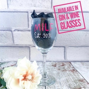 May include: A clear glass wine glass with the text "MILF Est. 2021" in pink lettering. The glass is sitting on a wooden surface with a white flower in the foreground. A pink square with the text "AVAILABLE IN GIN & WINE GLASSES" is in the background.