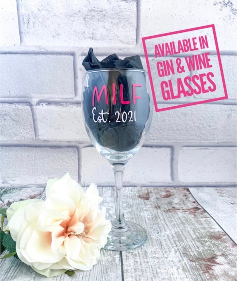 May include: A clear glass wine glass with the text "MILF Est. 2021" in pink lettering. The glass is sitting on a wooden surface with a white flower in the foreground. A pink square with the text "AVAILABLE IN GIN & WINE GLASSES" is in the background.