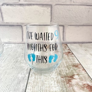 May include: A clear glass wine glass with the text "I've waited 9 months for this" and blue baby footprints.