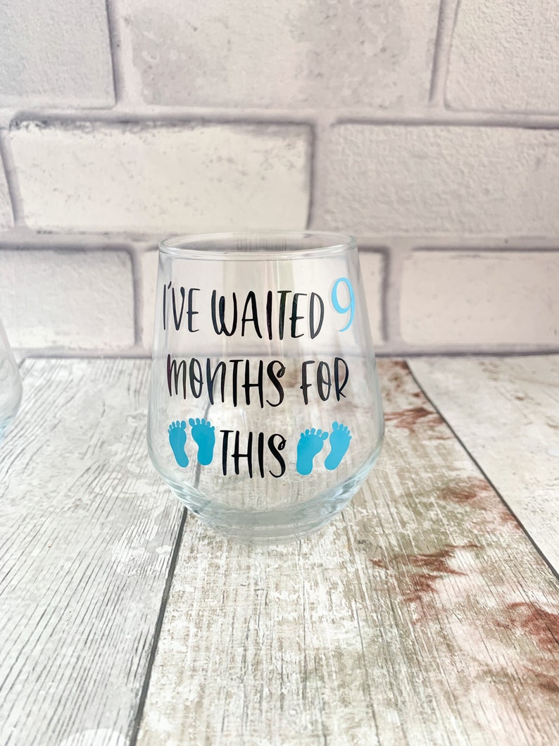 May include: A clear glass wine glass with the text "I've waited 9 months for this" and blue baby footprints.