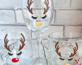 May include: Three glass mugs with reindeer faces on them. The mugs are clear glass and have brown antlers, black eyelashes, and a gold nose. One mug has a red nose and the name "Joana" written on it. Another mug has a gold nose and the name "Joe" written on it.