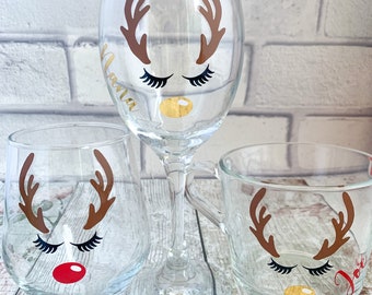 May include: Three clear glasses with reindeer face designs. The glasses include a wine glass, a stemless wine glass, and a mug. Each glass features brown antlers, black eyelashes, and a gold or red nose. The wine glass has the name "Nana" in gold.