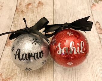 May include: Two personalised Christmas baubles. One is silver glitter with the name "Aarav" and snowflake accents. The other is red glitter with the name "Sahil" and snowflake accents. Both baubles have black ribbon bows.