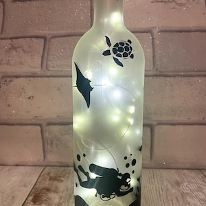 Könnte beinhalten: Eine gefrostete Glasflasche mit einer Lichterkette aus weißen Lichtern im Inneren. Die Flasche ist mit schwarzen Silhouetten eines Tauchers, eines Mantarochens, einer Meeresschildkröte und Blasen verziert.