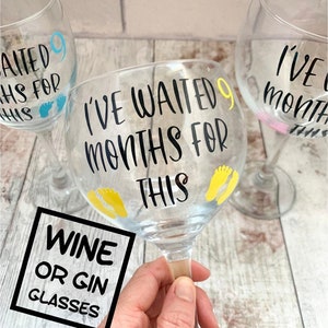 May include: Two clear glass wine or gin glasses with black vinyl lettering that reads "I've waited 9 months for this". One glass has blue footprints and the other has yellow footprints.