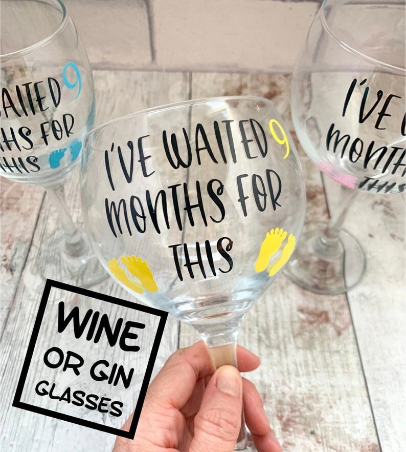 May include: Two clear glass wine or gin glasses with black vinyl lettering that reads "I've waited 9 months for this". One glass has blue footprints and the other has yellow footprints.