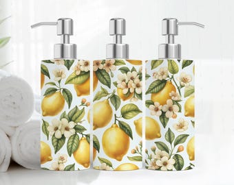 May include: Three decorative soap dispensers with a stainless steel pump. The dispensers feature a white background with a pattern of yellow lemons, green leaves, and white flowers. The dispensers are cylindrical in shape.