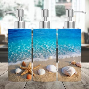 May include: Three silver soap dispensers with a beach scene design. The dispensers feature a blue ocean with white waves and seashells on a sandy beach.