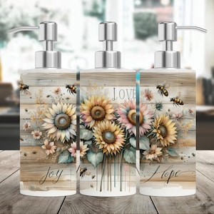 May include: Set of three ceramic soap dispensers with a rustic woodgrain background and sunflower and bee designs. The dispensers have the words "Joy", "Love", and "Hope" printed on them.