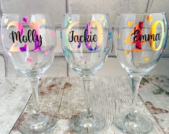 May include: Three clear wine glasses with iridescent numbers and names on them. The glasses are decorated with small hearts and butterflies. The names on the glasses are Molly 21, Jackie 60, and Emma 40.