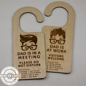 May include: Two wooden door hangers with a cartoon illustration of a person wearing glasses. The top hanger says "Dad is in a meeting, please do not disturb". The bottom hanger says "Dad is at work, visitors welcome".