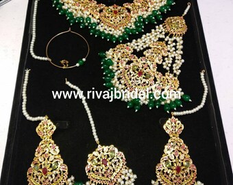 Op de afbeelding: Een traditionele Indiase bruidssieradenset met een gouden ketting, oorbellen, een maang tikka en een neusring. De set is versierd met parels, groene kralen en rode en roze stenen.