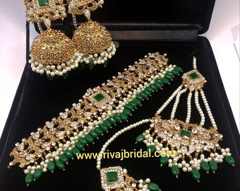 Op de afbeelding: Een goudkleurige bruidssieradenset met smaragdgroene accenten en witte parels. De set bevat oorbellen, een ketting, een hoofdband en een tikka. De sieraden worden gepresenteerd op een zwart oppervlak. De website www.rivajbridal.com is zichtbaar.
