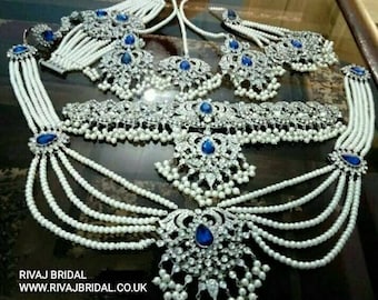 Op de afbeelding: Een zilveren bruidsjuwelenset met blauwe en witte stenen. De set bevat een ketting, oorbellen, een maang tikka en een tailleketting. De juwelen zijn versierd met parels en ingewikkelde ontwerpen.