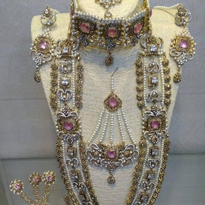 Peut inclure: Un ensemble de bijoux de mariée indien traditionnel avec une finition or et argent. L'ensemble comprend un collier ras du cou, un long collier, des boucles d'oreilles, un maang tikka et un ensemble de bracelets. Les bijoux sont ornés de pierres roses et blanches.