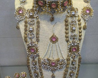 May include: A traditional Indian bridal jewellery set with a gold and silver finish. The set includes a choker necklace, a long necklace, earrings, a maang tikka, and a set of bangles. The jewellery is adorned with pink and white stones.