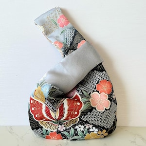 May include: A Japanese-style knot bag featuring a black fabric base with a vibrant floral pattern in shades of red, pink, and yellow. The bag has a solid light gray handle and interior lining, with a distinct asymmetrical design where one handle loops through the other.