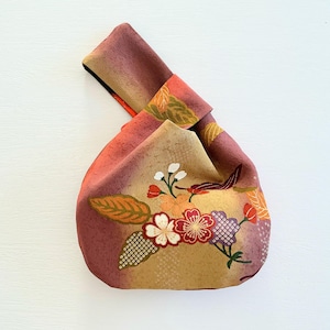 May include: A Japanese-style knot bag featuring a gradient of muted red, orange, and beige tones. The fabric is decorated with a floral pattern including white, red, and purple blossoms, green leaves, and a subtle geometric texture throughout.