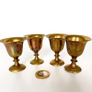 Pode incluir: Conjunto de quatro pequenas taças de latão dourado com desenhos gravados intrincados. As taças têm uma base arredondada e uma parte superior alargada.