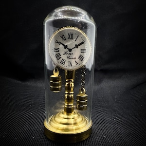 May include: A small, antique-style clock with a gold-toned finish and Roman numeral face. The clock is encased in a glass dome and sits on a gold-toned base.