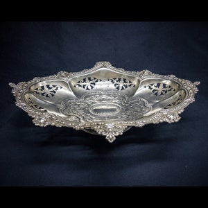 May include: An ornate silver-coloured serving dish with a decorative, pierced design. The oval-shaped dish features intricate floral and scrollwork patterns, with a raised, scalloped edge. The dish is supported by a small, decorative base, against a dark background.