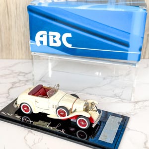 以下が含まれることがあります： クリーム色のヴィンテージコンバーチブルモデルカーで、深紅のインテリアと赤いスポークホイールが特徴です。車は銀色のプレートが付いた黒いディスプレイベースに固定されており、ABCと書かれた青い箱と透明なアクリル製のディスプレイケースの前に置かれています。