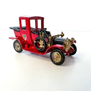 Op de afbeelding: Een rode speelgoedauto met zwarte wielen en gouden accenten. De auto heeft een zwart interieur en een witte achtergrond.