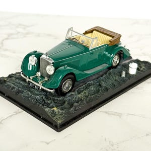 Könnte beinhalten: Ein grünes Oldtimer-Cabrio-Modell mit beigefarbenem Innenraum und braunem Verdeck. Das Modell steht auf einer schwarzen Basis mit strukturierter Oberfläche. Das Nummernschild lautet "YFF 417".