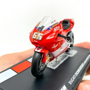 Może przedstawiać: Model motocykla Ducati Desmosedici w kolorze czerwonym i białym z numerem 65 i napisem "Alice" na boku. Model stoi na czarnej podstawie z logo szachownicy.