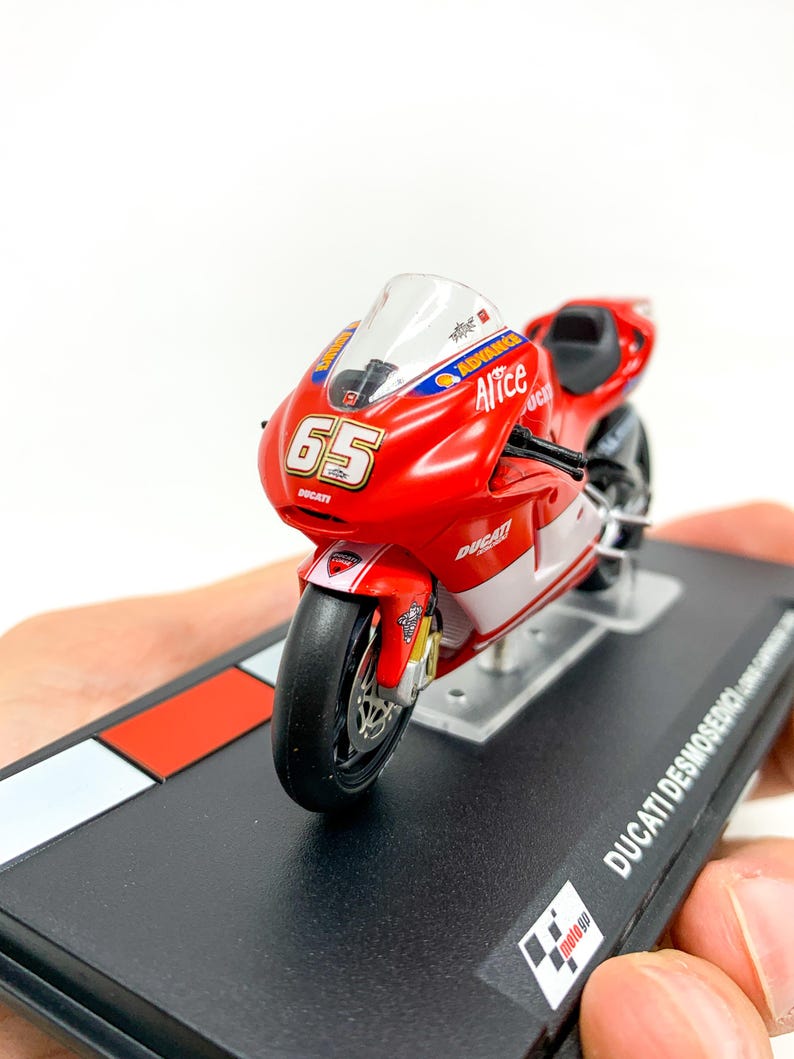 Może przedstawiać: Model motocykla Ducati Desmosedici w kolorze czerwonym i białym z numerem 65 i napisem "Alice" na boku. Model stoi na czarnej podstawie z logo szachownicy.