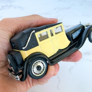 May include: A black and yellow vintage toy car model with a black roof and chrome accents. The car is in a hand.
