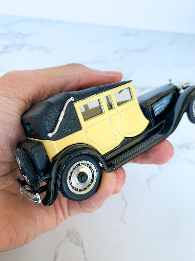 May include: A black and yellow vintage toy car model with a black roof and chrome accents. The car is in a hand.