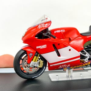 Może przedstawiać: Model motocykla Ducati Desmosedici w kolorze czerwonym i białym z napisem "Alice" na boku. Motocykl stoi na przezroczystym stojaku z czarną podstawą, na kt&oacute;rej widnieje napis "Ducati Desmosedici".