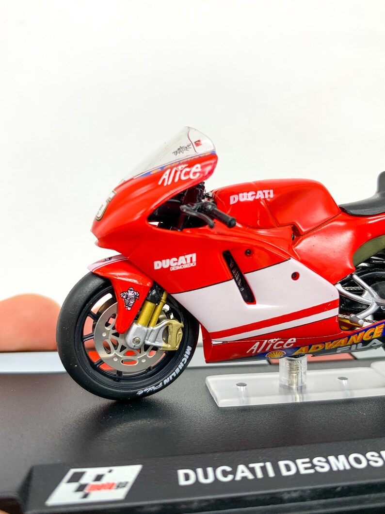 Może przedstawiać: Model motocykla Ducati Desmosedici w kolorze czerwonym i białym z napisem "Alice" na boku. Motocykl stoi na przezroczystym stojaku z czarną podstawą, na kt&oacute;rej widnieje napis "Ducati Desmosedici".