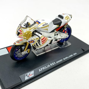 Peut inclure: Modèle de moto Aprilia RS3 blanc et or avec le numéro 99 sur le côté. Le modèle est sur un socle d'exposition noir avec le texte "APRILIA RS3 JEREMY MCWILLIAMS 2004".