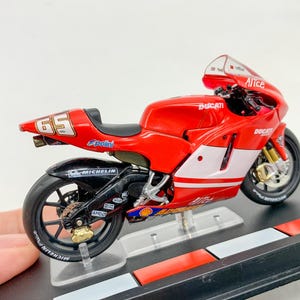 Może przedstawiać: Model motocykla Ducati w kolorze czerwonym i białym z numerem 65 na boku. Motocykl stoi na czarnym stojaku z czerwono-białym wzorem w kratkę.