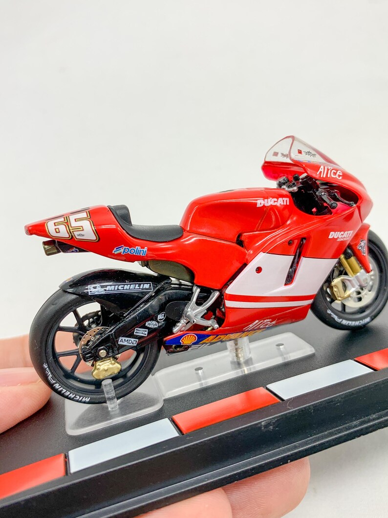 Może przedstawiać: Model motocykla Ducati w kolorze czerwonym i białym z numerem 65 na boku. Motocykl stoi na czarnym stojaku z czerwono-białym wzorem w kratkę.