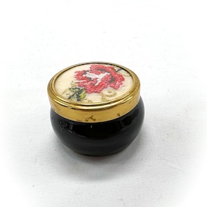 May include: A small, round, black glass jar with a gold rim and a white lid featuring a red embroidered flower.