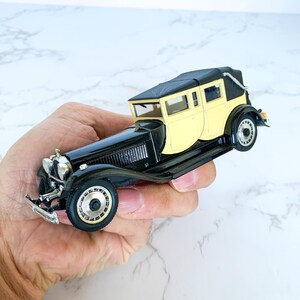 May include: A black and cream vintage toy car model with a black fabric roof. The car has a detailed interior and chrome accents.
