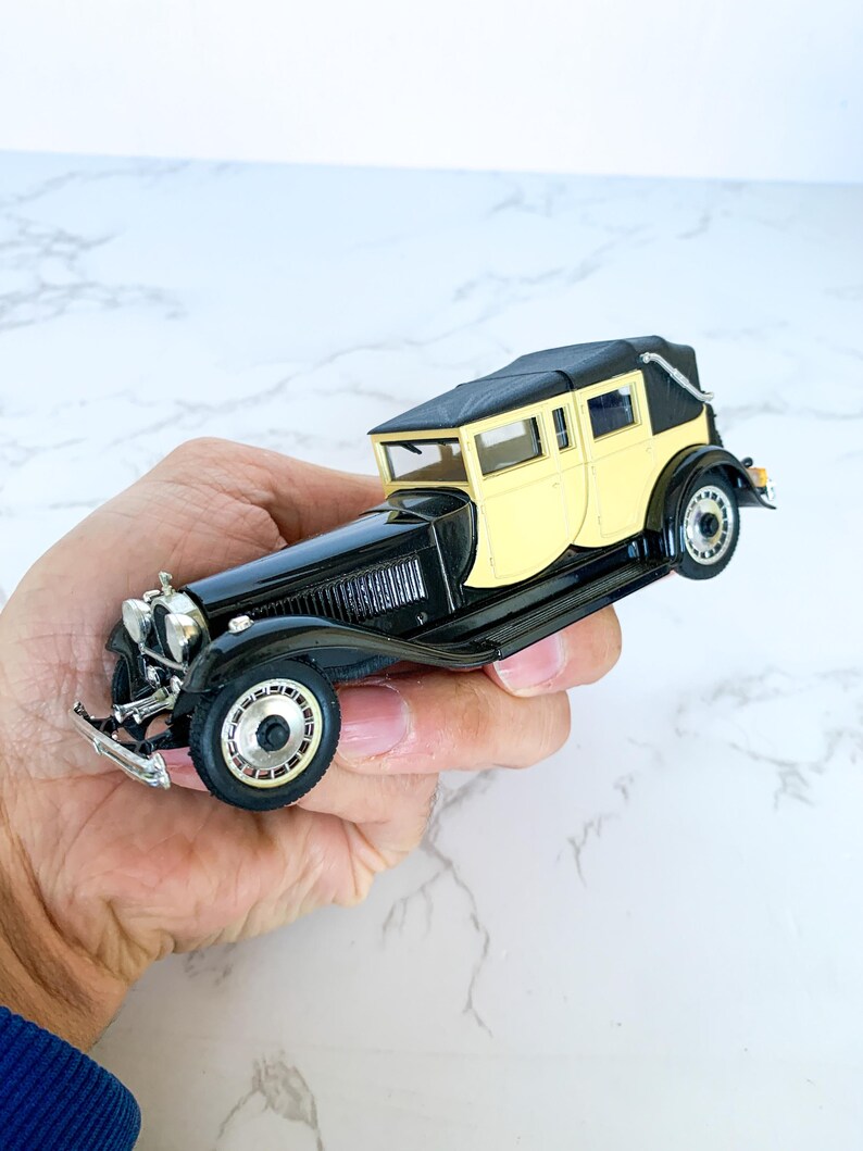 May include: A black and cream vintage toy car model with a black fabric roof. The car has a detailed interior and chrome accents.